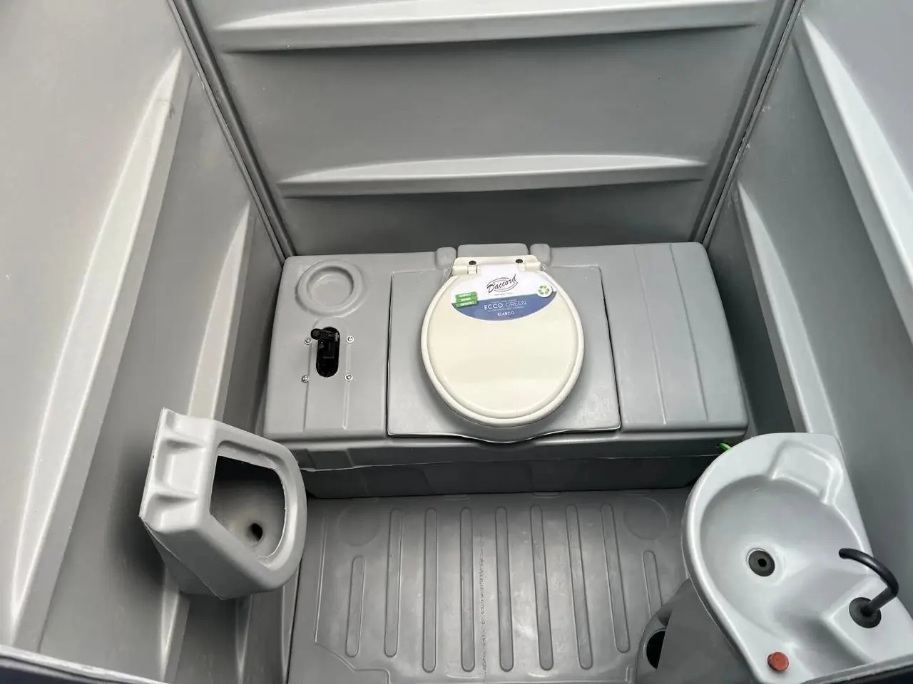 Interior de un baño químico visto desde arriba: inodoro con tapa, mingitorio y lavamanos
