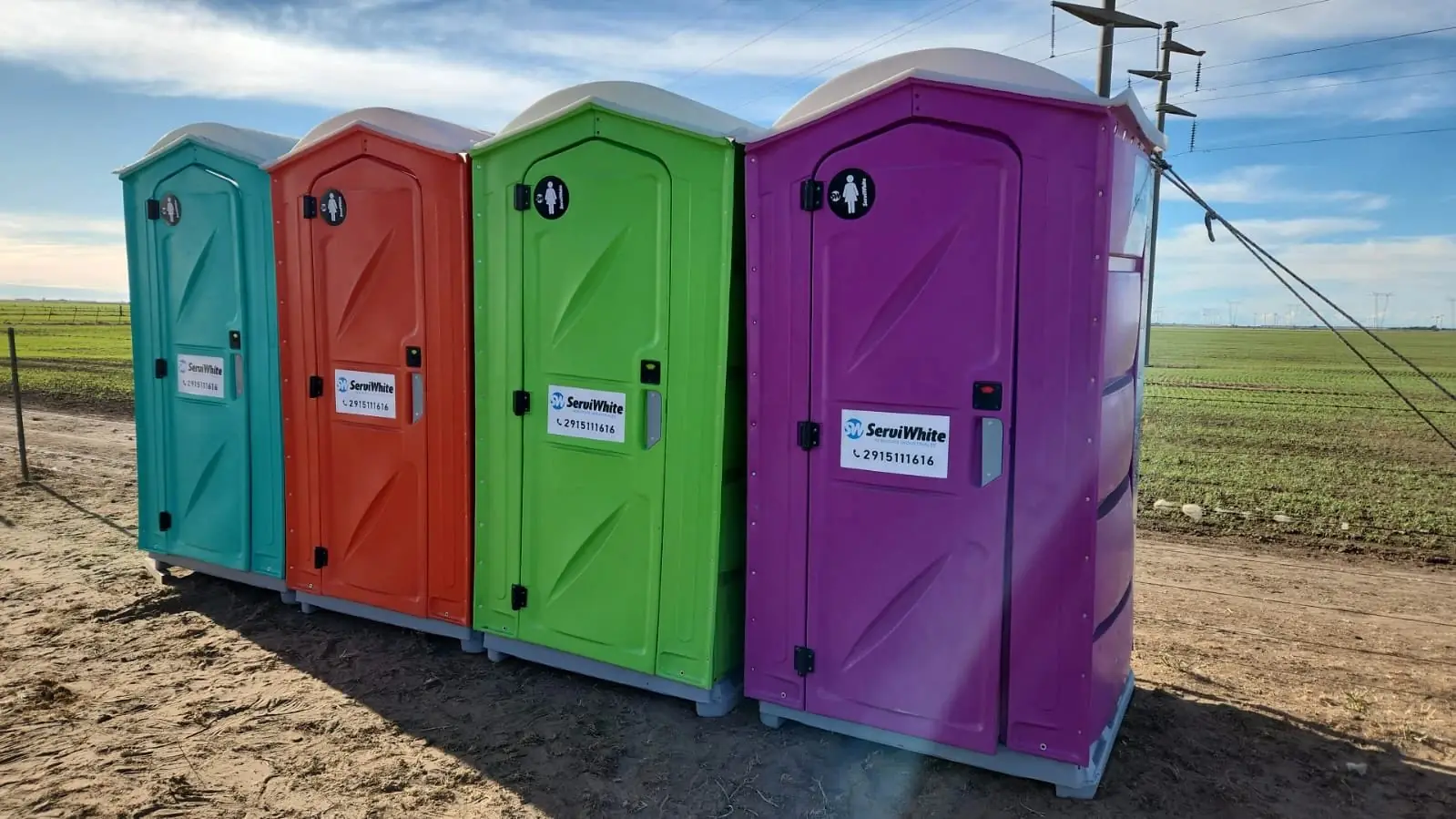 Cuatro baños químicos Serviwhite turquesa, naranja, verde y violeta instalados en un campo