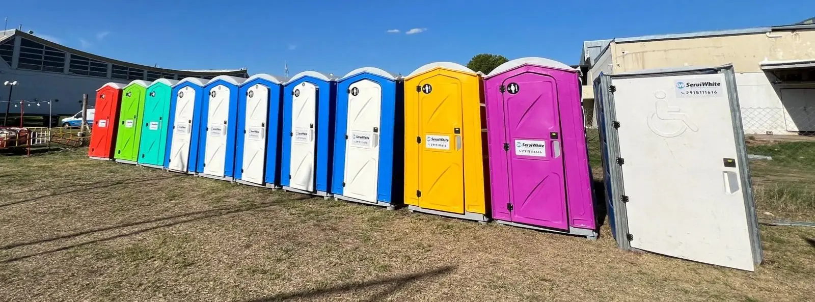 Hilera de baños químicos Serviwhite de colores en un evento, con una cabina accesible para personas con discapacidad