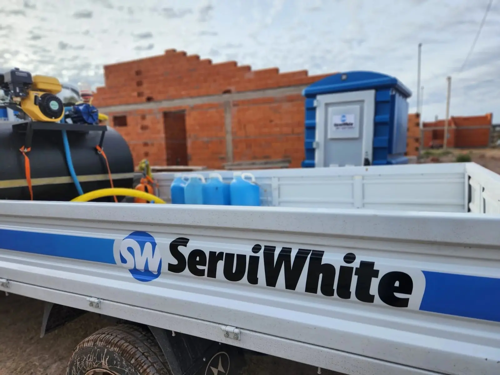 Camioneta Serviwhite con tanque de limpieza y bidones de desinfectante junto a un baño químico en obra