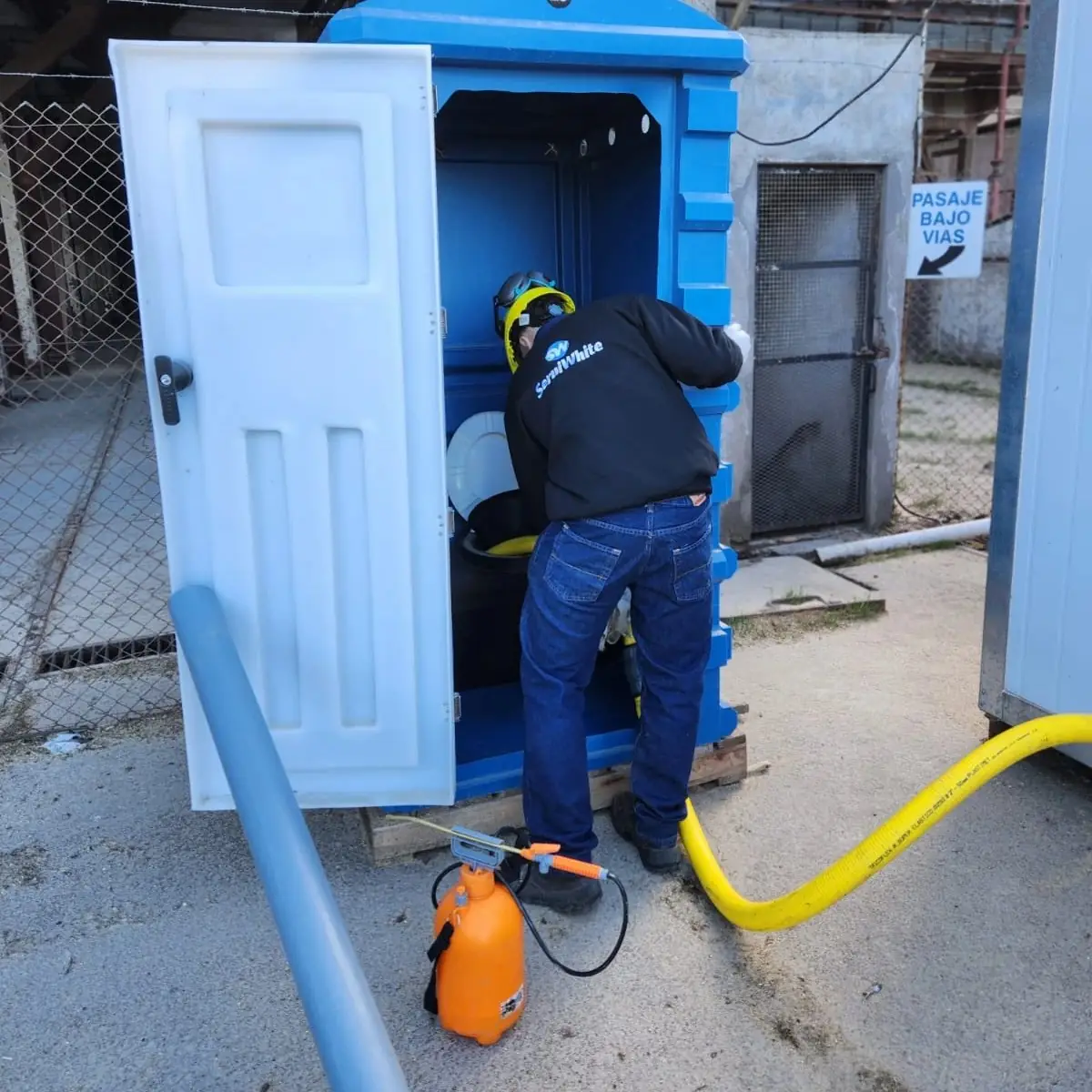 Operario de Serviwhite con casco limpia y desinfecta un baño químico con manguera de succión