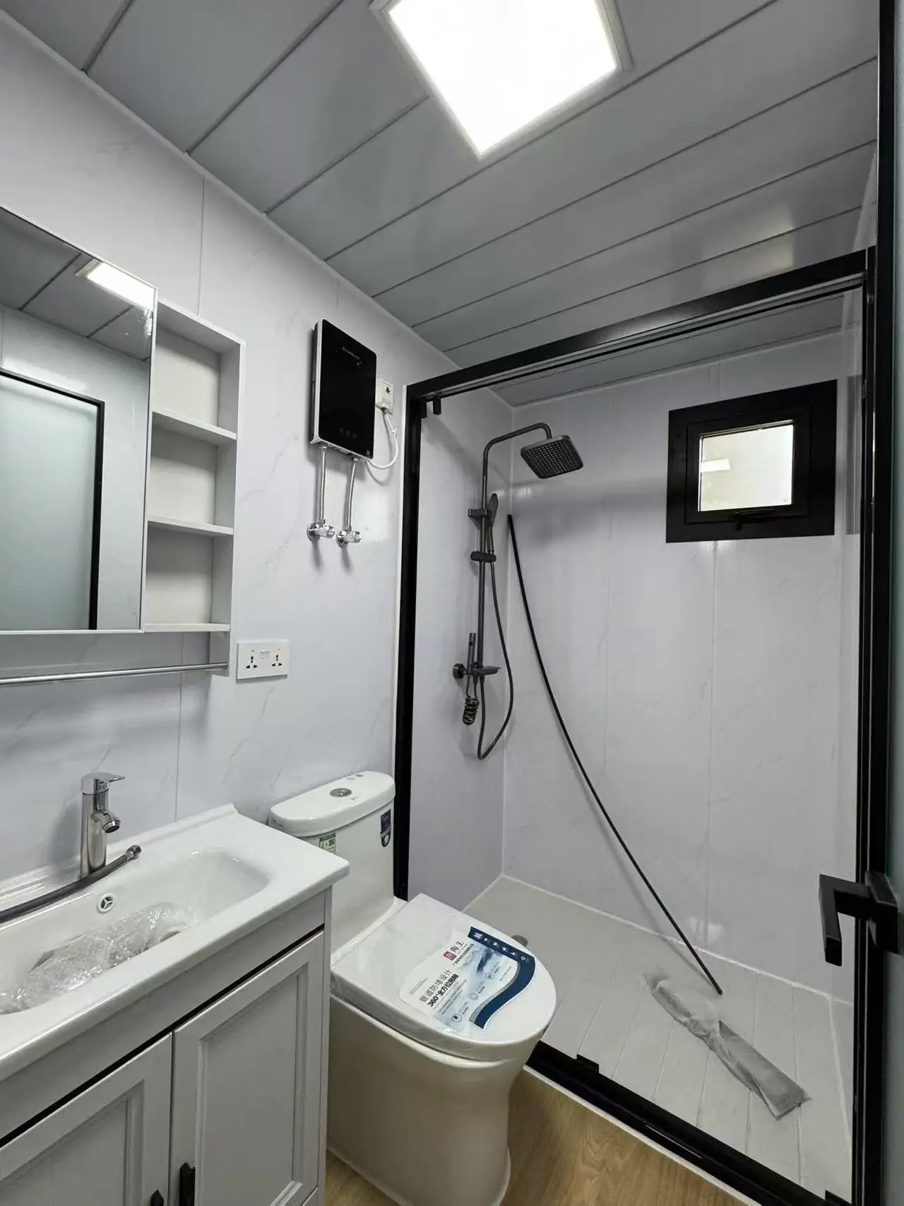 Baño de una casa expandible con ducha de mampara, ventana, inodoro, vanitory con espejo y calefón eléctrico