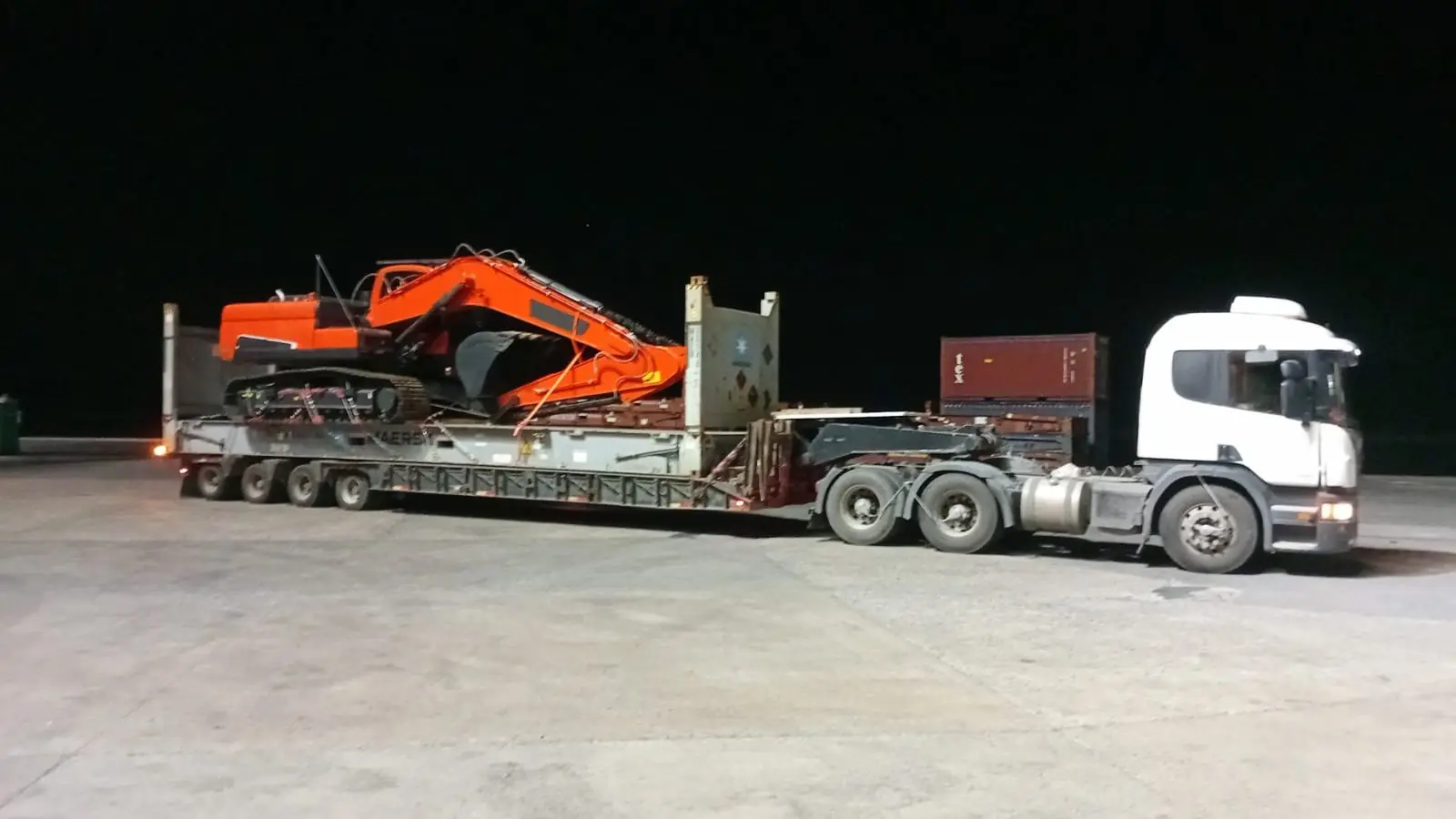 Excavadora Doosan naranja sobre carretón de cuatro ejes, de noche en el puerto