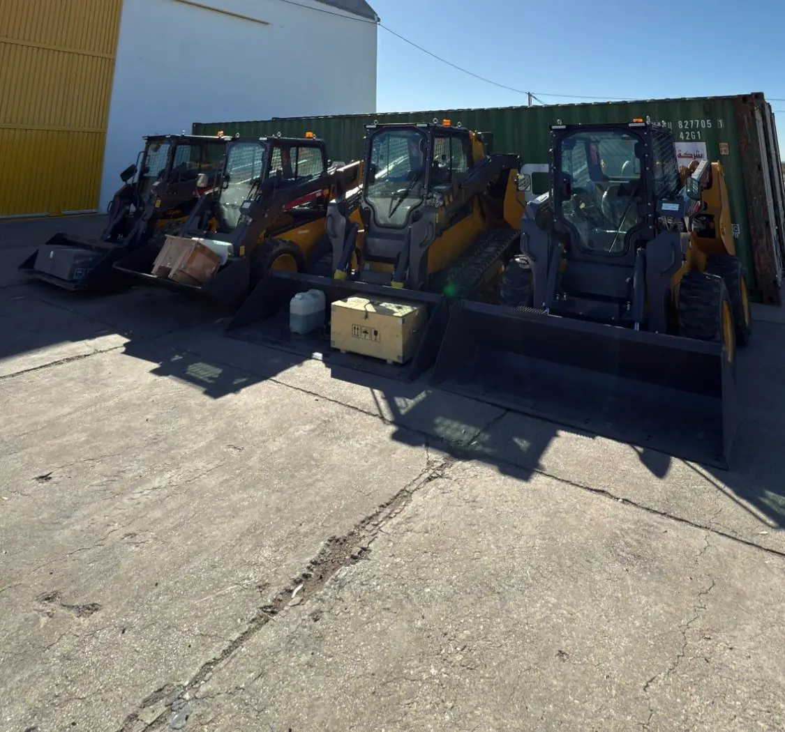 Cuatro minicargadoras XCMG con balde, alineadas al sol frente a un galpón y un contenedor