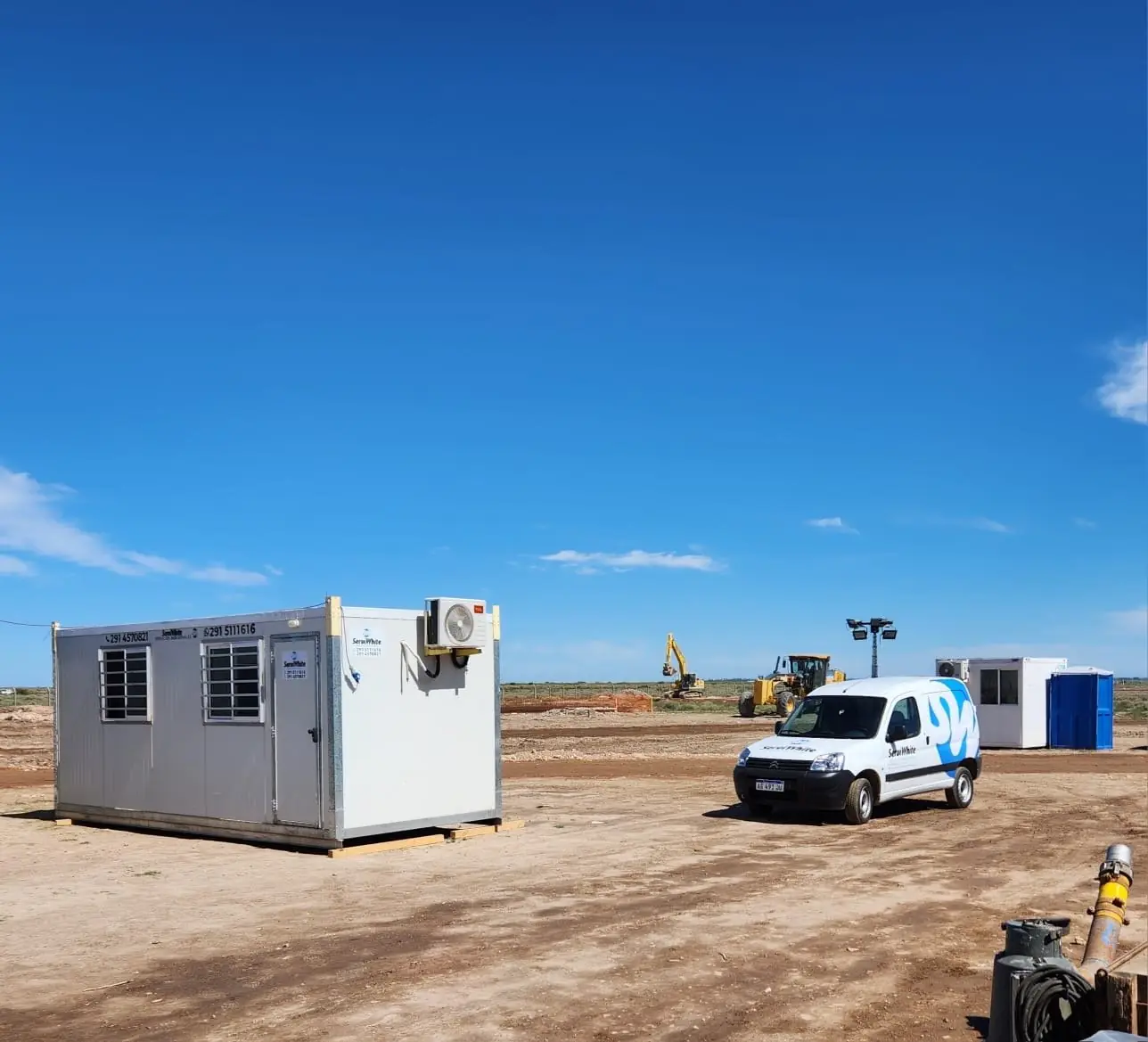 Módulo oficina de Serviwhite con aire acondicionado en una obra, con la camioneta de la empresa, una torre de iluminación y un baño químico