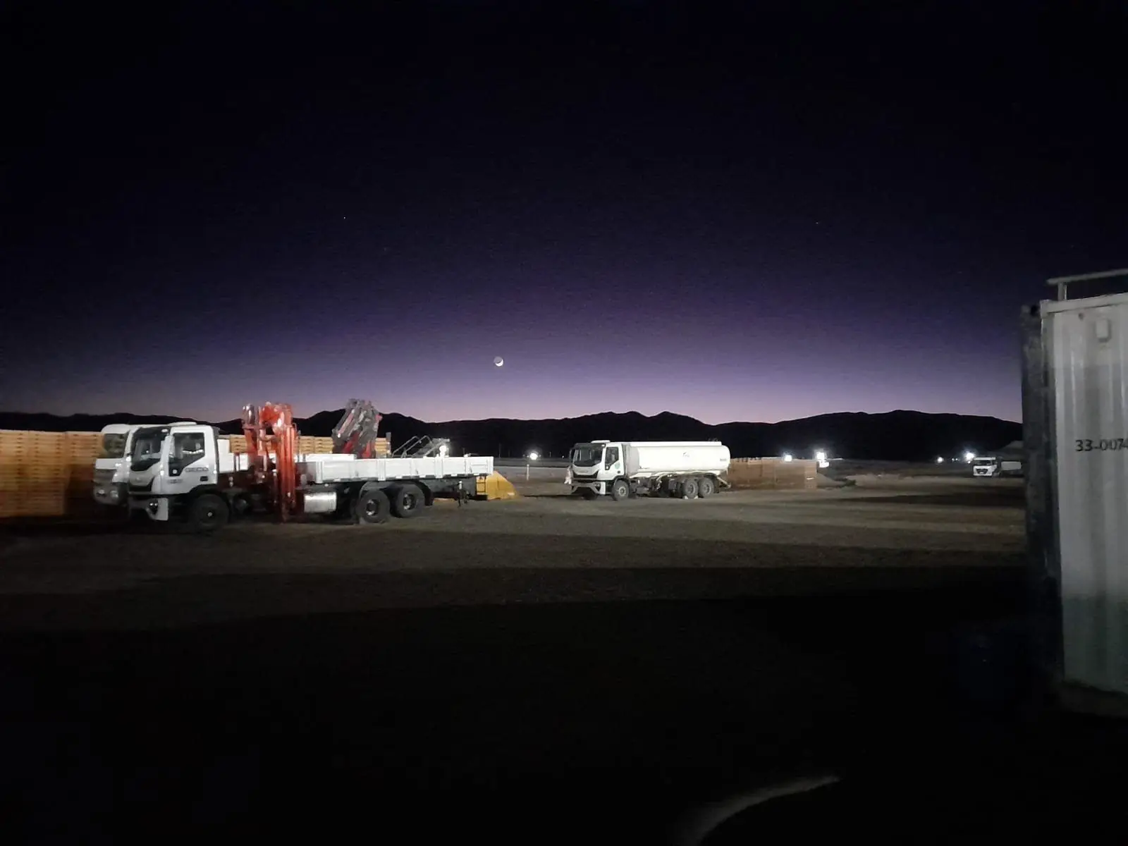 Camión con hidrogrúa y camión cisterna en un predio al anochecer, con cerros de fondo