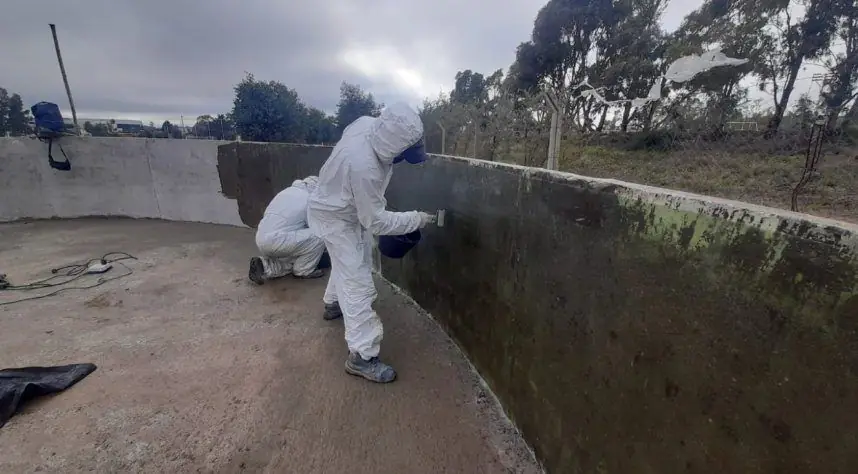 Dos operarios con traje descartable impermeabilizan con pincel la pared de una cisterna de hormigón