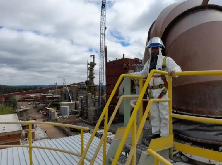 Operario con traje descartable, arnés y careta en una plataforma elevada sobre una planta industrial